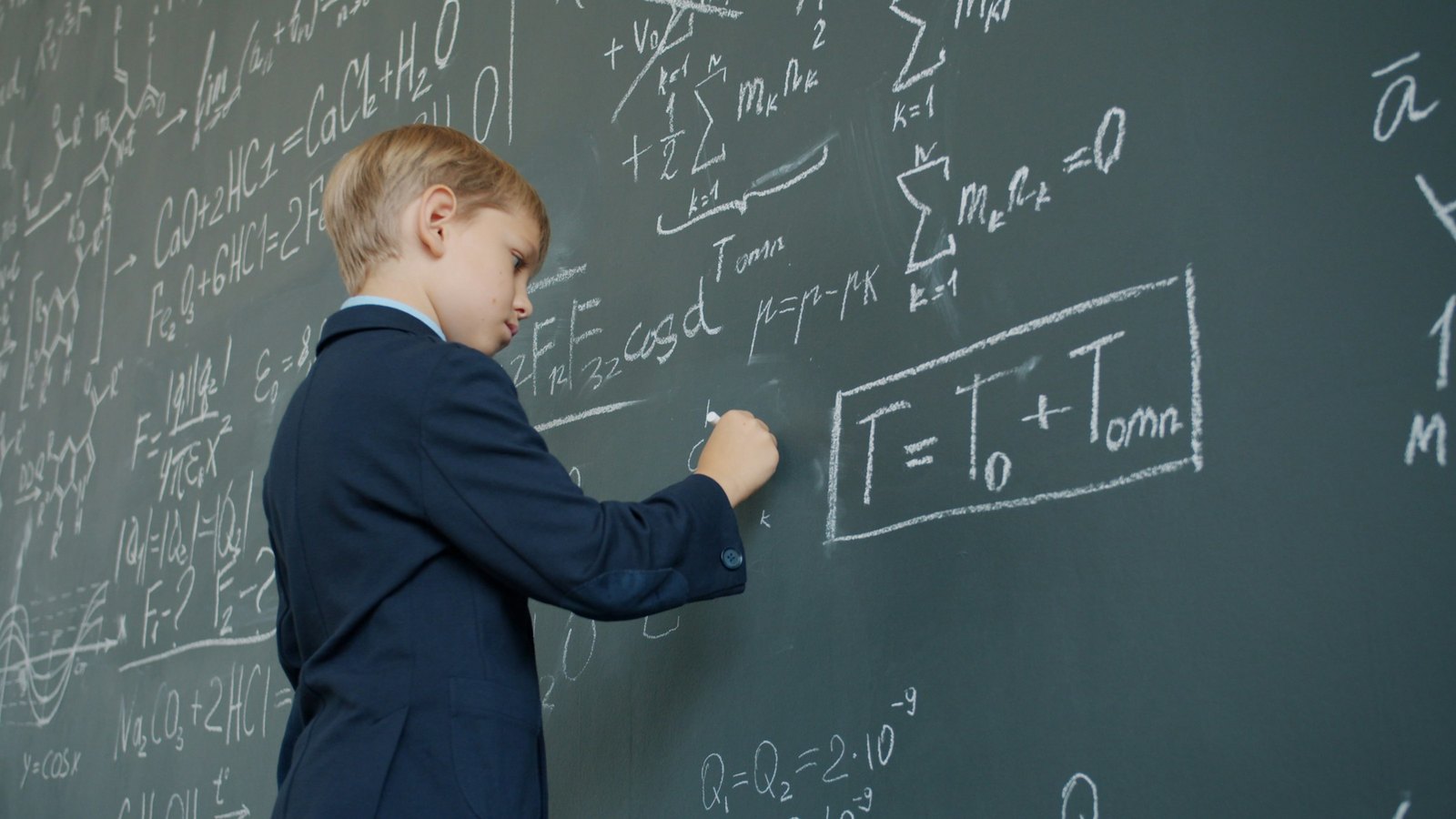 Anak laki-laki berjas menulis rumus matematika di papan tulis kapur
