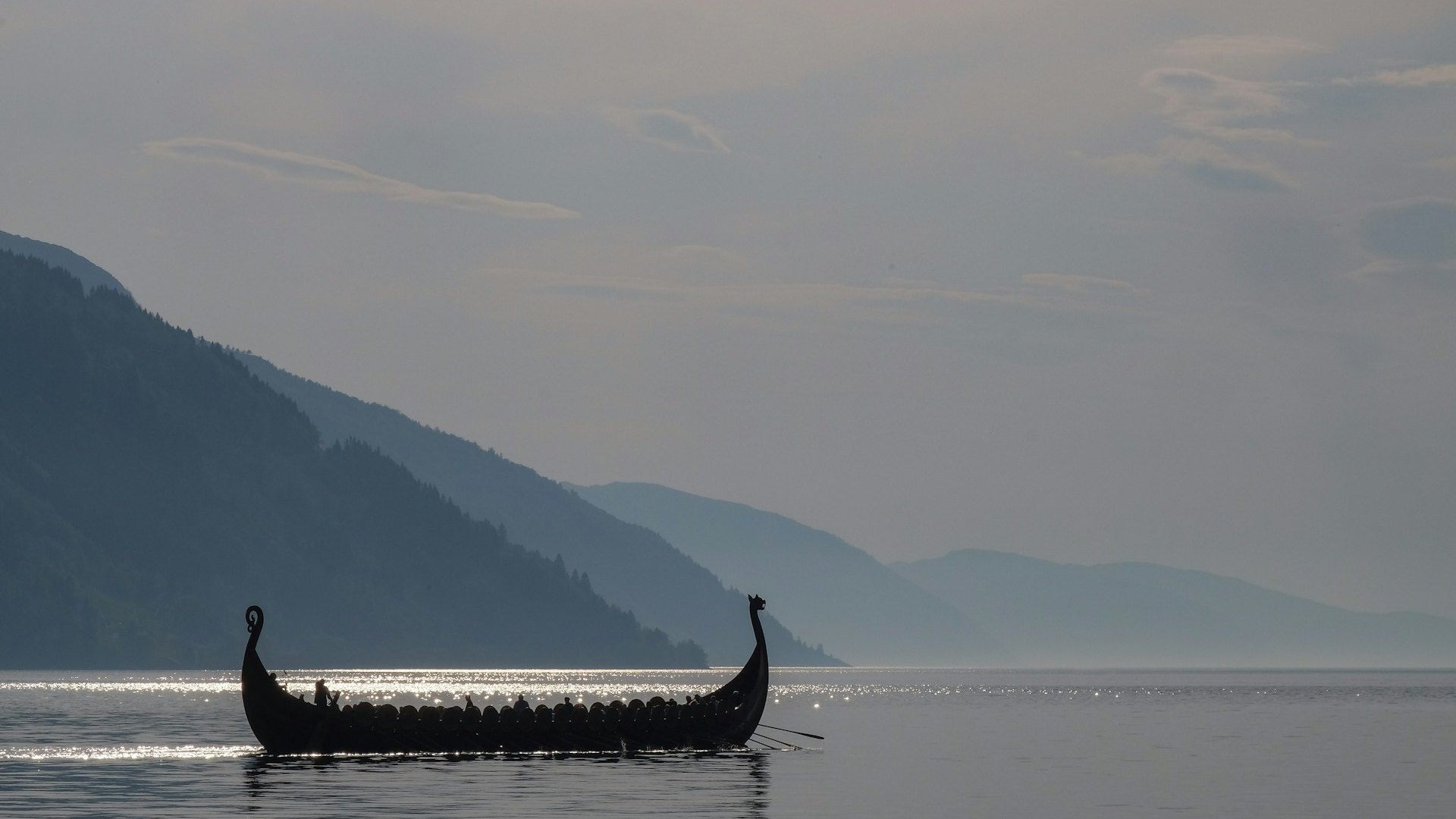 Siluet replika kapal panjang Viking berlayar di fjord dengan latar pegunungan berkabut