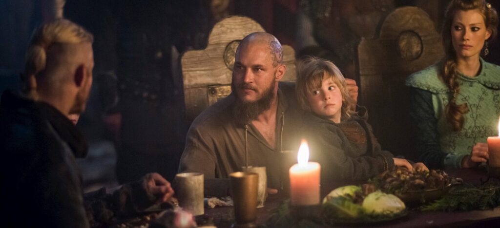 Travis Fimmel sebagai Ragnar Lothbrok duduk di meja perjamuan bercahaya lilin sambil merangkul seorang anak dalam serial Vikings